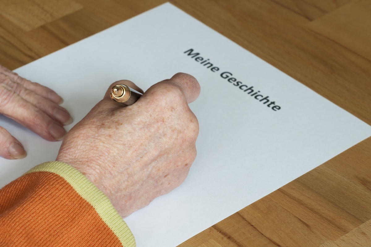 Ältere Hand hält einen Stift und schreibt auf ein Blatt Papier mit der Aufschrift Meine Geschichte